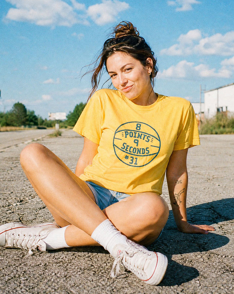 Woman sitting on a road wearing a yellow t-shirt with text, blue shorts, and white sneakers.