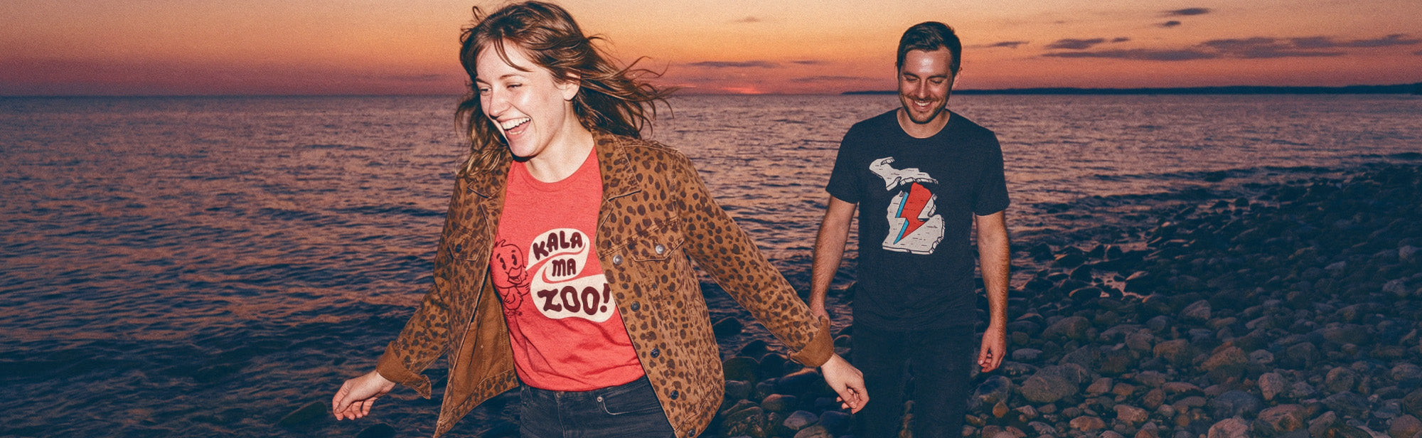 Two people walking on a beach at sunset wearing Michigan themed HomeTown Riot apparel