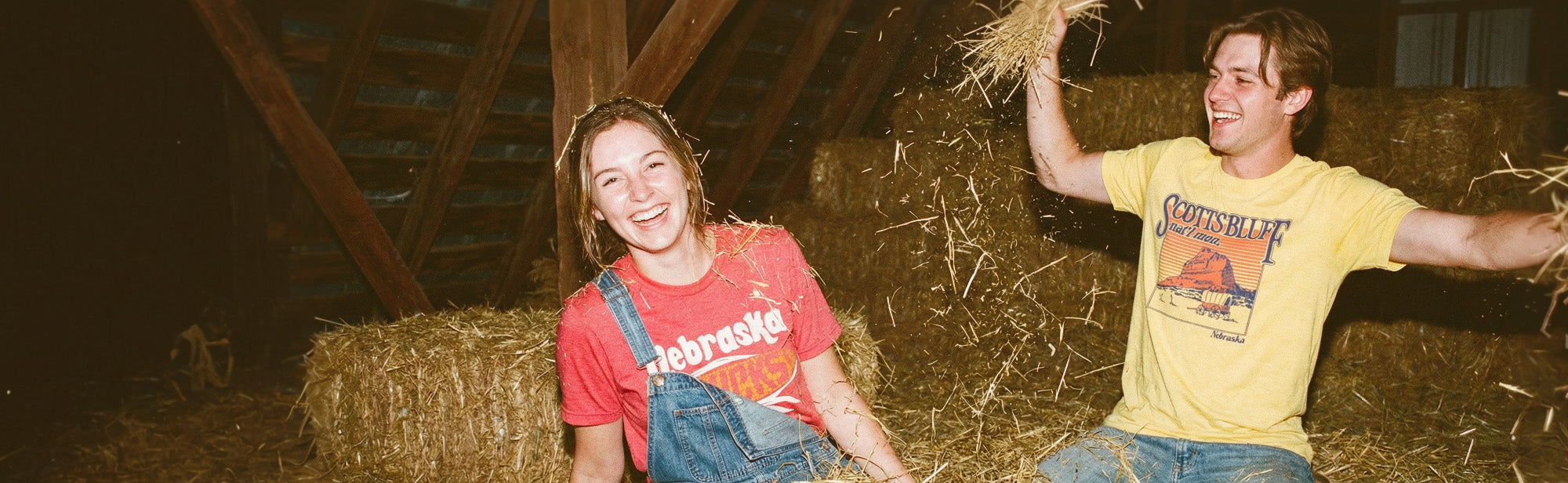 Two people wearing Nebraska themed HomeTown Riot shirts in a hay loft of a barn