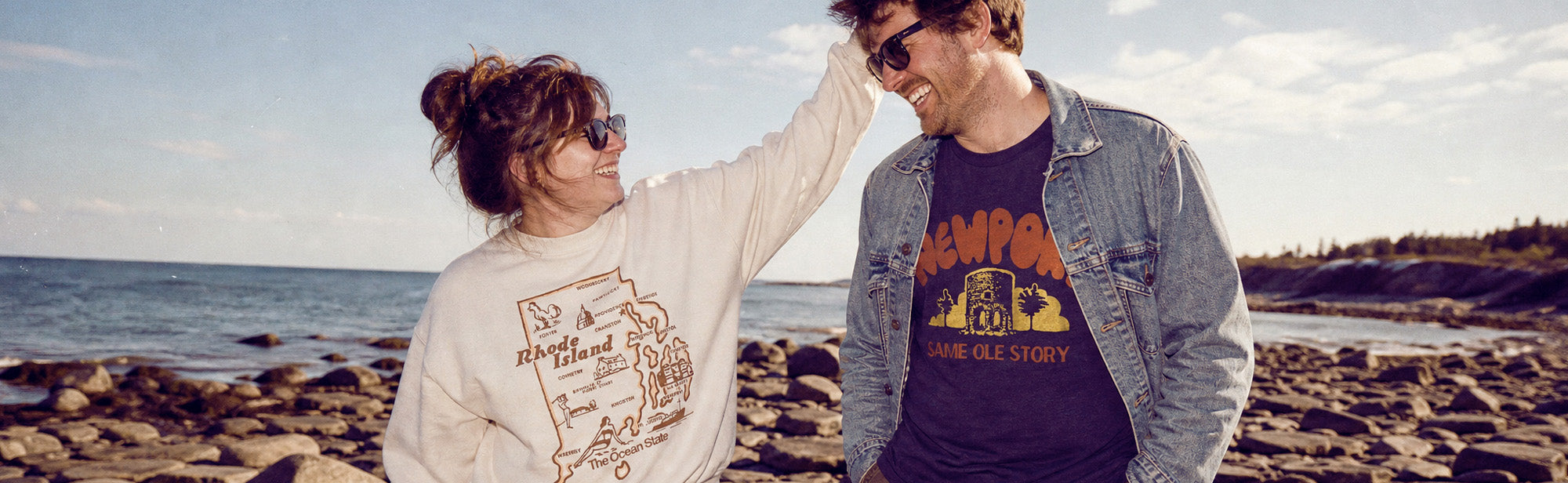 A couple standing on a rocky beach with the ocean in the background. The woman wears a cream sweatshirt with a graphic print and sunglasses, and the man wears a denim jacket over a dark graphic t-shirt.