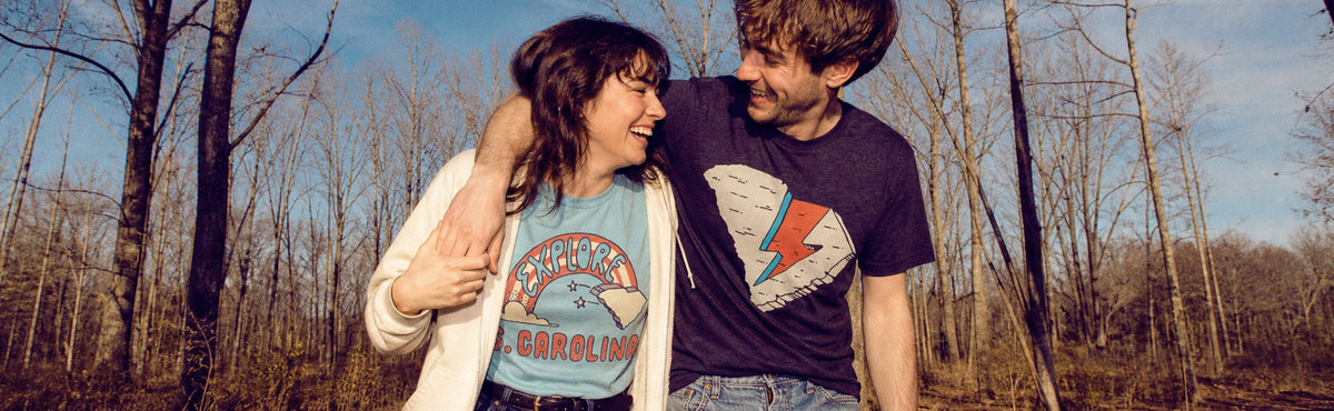 A couple outdoors in a wooded area, with the man wearing a dark graphic T-shirt featuring a white wing and red lightning bolt, and the woman wearing a light blue graphic T-shirt with 'North Carolina' text.