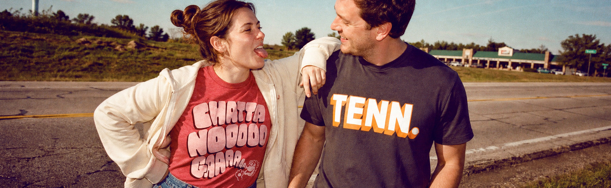 Two people standing outdoors on a roadside, each wearing a graphic T-shirt. The person on the left wears a red T-shirt with large white text reading 'CHAT! NODOO! CHAT!' and a white jacket, while the person on the right wears a black T-shirt with large orange and white text reading 'TENN.