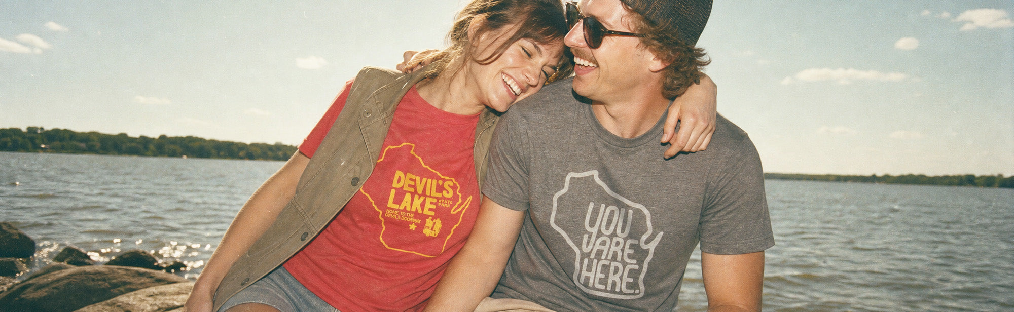 A couple sitting together outdoors by a lake, wearing graphic t-shirts. The woman wears a red t-shirt with a yellow 'Devil's Lake' graphic, and the man wears a gray t-shirt with a white 'UDY VACANCY' graphic.