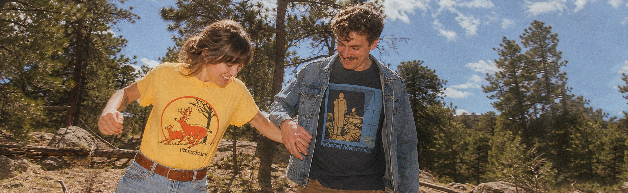 Two people outdoors in a pine forest, each wearing a graphic T-shirt. The person on the left wears a yellow T-shirt with a deer graphic, and the person on the right wears a blue denim jacket over a dark gray T-shirt with a cityscape graphic.